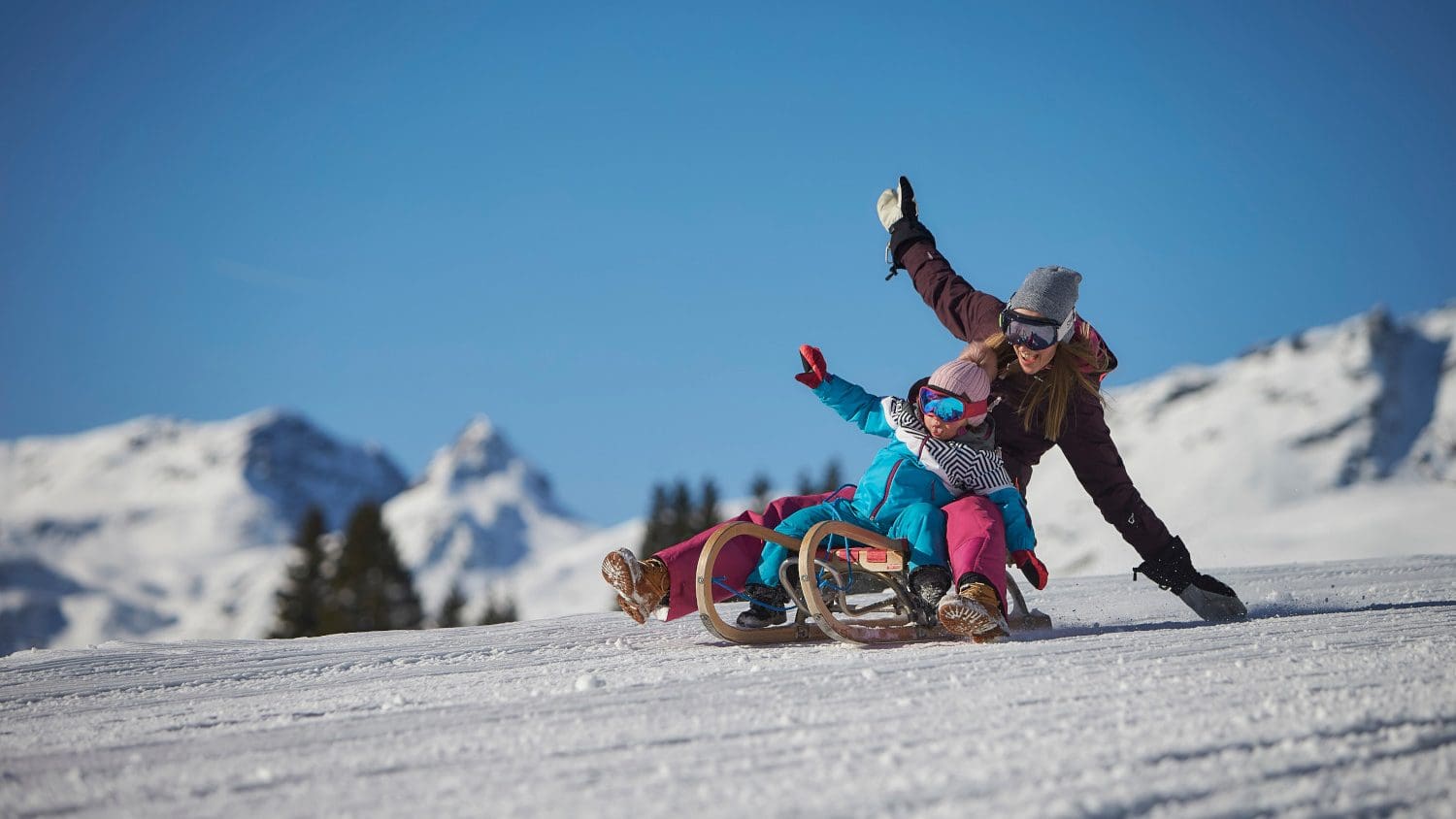 Frau und Kind rodeln bei Sonne und blauem Himmel eine verschneite Piste in Saalbach-Hinterglemm  hinab.