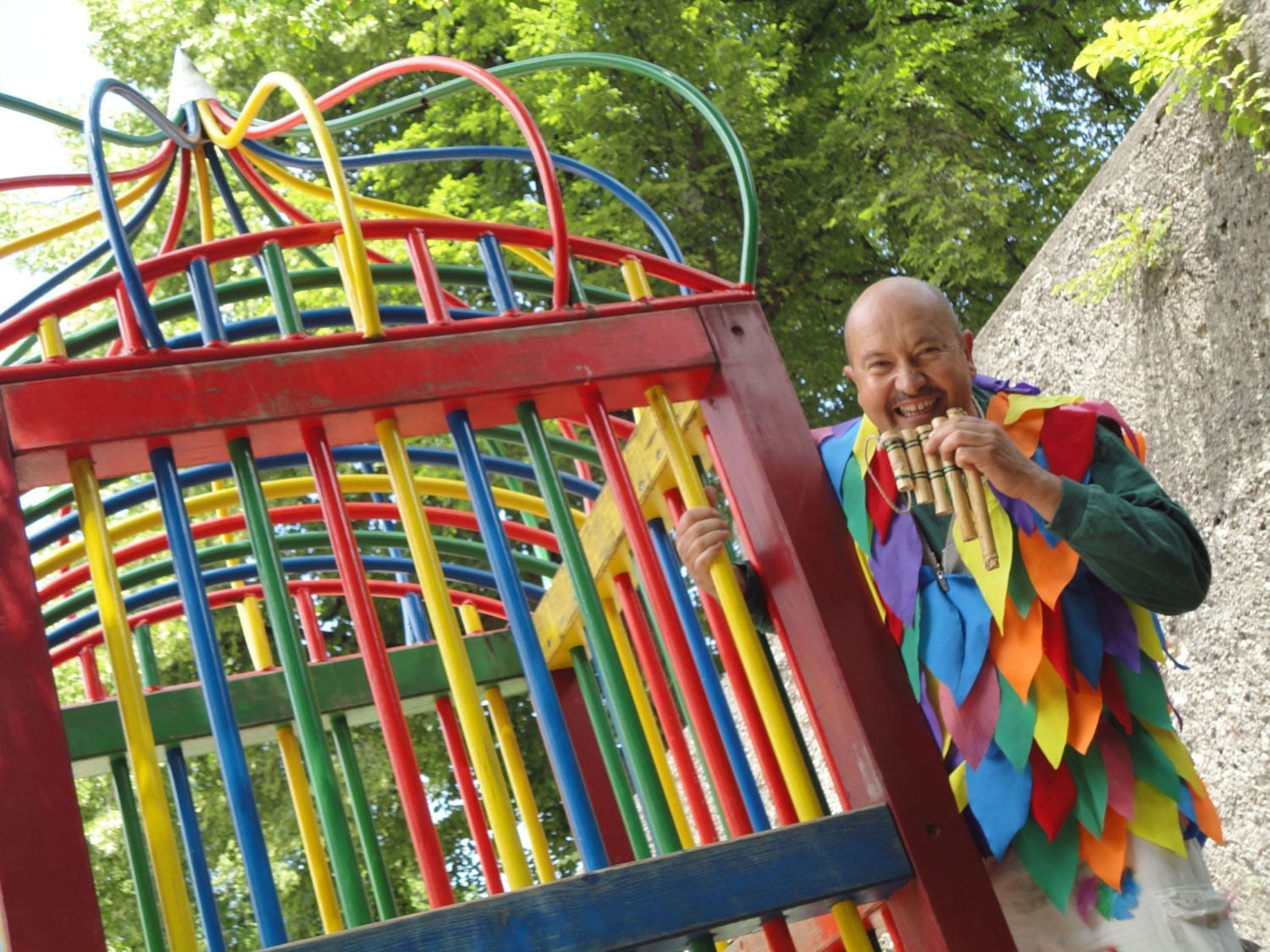 Der Kindermusical-Darsteller Manfred Schwaiger hält sich an einem bunten Kinderklettergerüst fest, während er ein buntes Kostüm trägt und eine Panflöte in der Hand hält.