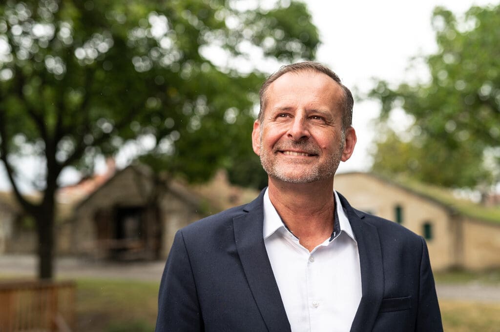 Martin Horak lächelnd im Porträt mit hellem Hemd und dunkel Sakko. Im Hintergrund erkennt man verschwommen die Kellergasse in Purbach.